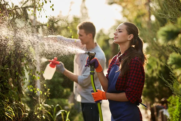 Trabaja en el jardín. Chica jardinero rocía agua y un chico rocía fertilizante en las plantas en el hermoso vivero-jardín en un día soleado . — Foto de Stock