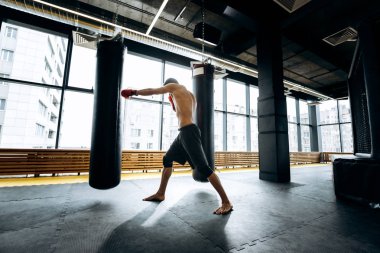 Siyah şort giymiş çıplak bir gövde ile kırmızı boks eldivenleri adam panoramik pencerelerin arka planına karşı spor salonunda yumruk torbası vurdu