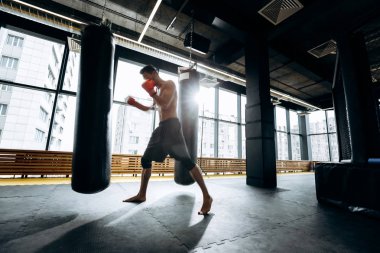 Siyah şort giymiş çıplak bir gövde ile kırmızı boks eldivenleri adam panoramik pencerelerin arka planına karşı spor salonunda yumruk torbası vurdu