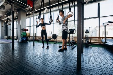 Siyah spor giysileri ve atletik adam giyinmiş ince koyu saçlı kız egzersiz ekipmanları yanında geniş spor salonunda konuşuyoruz