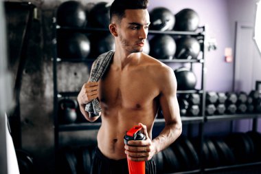 Çıplak bir gövde ile atletik adam omuzunda t-shirt tutar ve spor salonunda spor ekipmanları yakın elinde su ile şişe tutar