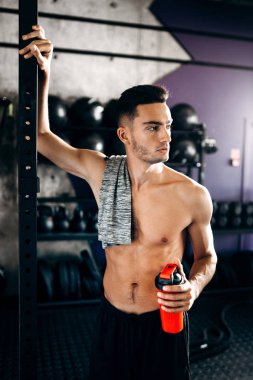 Çıplak bir gövde ile atletik adam omuzunda t-shirt tutar ve spor salonunda spor ekipmanları yakın elinde su ile şişe tutar