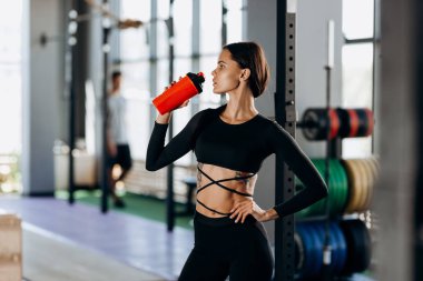 Siyah spor kıyafetleri giymiş ince koyu saçlı kız spor ekipmanlarının yakınındaki jimnastik salonunda su içiyor