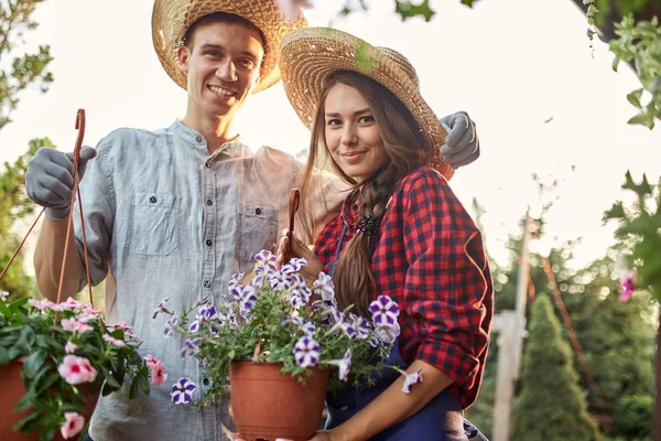 Jardineiros Guy e menina em um chapéu de palha segurar panelas com petúnia maravilhosa no caminho do jardim em um dia ensolarado . — Fotografia de Stock