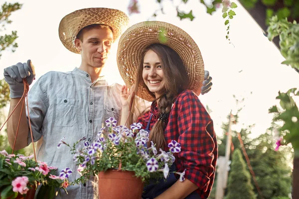 Jardineiros Guy e menina em um chapéu de palha segurar panelas com petúnia maravilhosa no caminho do jardim em um dia ensolarado . — Fotografia de Stock