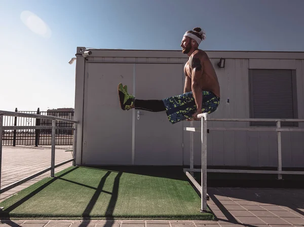 Jeune homme athlétique avec un torse nu avec des tatouages et un bandeau vêtu de leggings noirs et de shorts bleus fait des exercices de sport sur des clôtures métalliques par une chaude journée ensoleillée — Photo