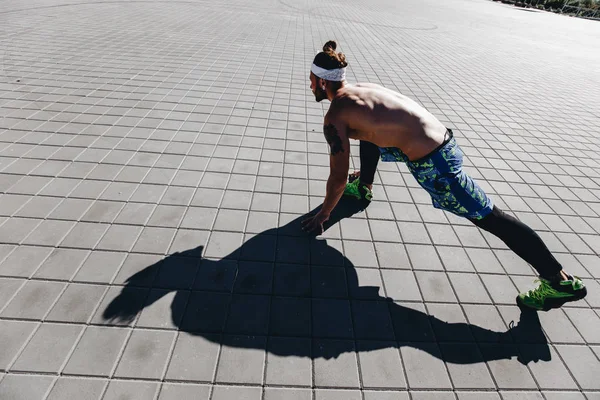 Jeune homme athlétique avec un torse nu avec des tatouages et un bandeau sur la tête habillé des leggings noirs et des shorts bleus commençant à courir sur les dalles de pavage sur la place par une chaude journée ensoleillée — Photo