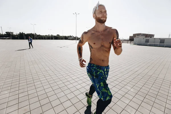 Homem atlético com um tronco nu com tatuagens e com a cabeça vestida com as pernas pretas e shorts azuis corre em lajes de pavimentação na praça em um dia ensolarado quente — Fotografia de Stock