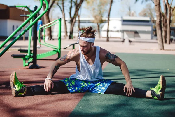 Athletic man with headband on his head and tattoos dressed in the white t-shirt, black leggings and blue shorts is doing stretching on the sports ground outside on a sunny day