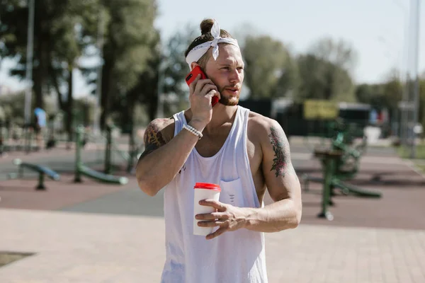 Young man with white headband dressed in a white t-shirt talked by phone and holds a plastic cup in his hand on the sports ground outside on a sunny day