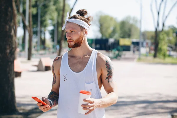 Jovem com a cabeça branca vestida com uma camiseta branca usando telefone celular e segura uma xícara de plástico na mão no chão esportivo do lado de fora em um dia ensolarado — Fotografia de Stock