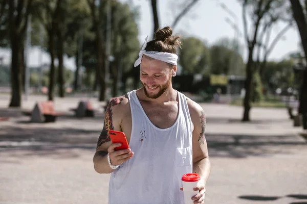 Smiling young man with white headband dressed in a white t-shirt using mobile phone and holds a plastic cup in his hand on the sports ground outside on a sunny day