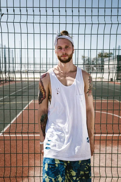 Young man with white headband  on his head and tattoos on his arms dressed in the white t-shirt, black leggings and blue shorts stands leaning on the playground fence outside on a sunny day