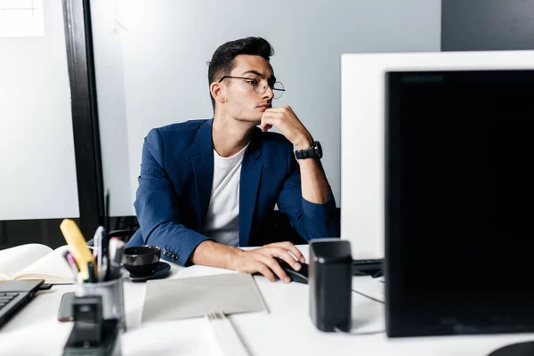 En ung mann med briller kledd i dress sitter ved en pc på kontoret. – stockfoto