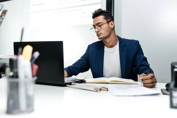 El joven arquitecto de cabello oscuro en gafas y chaqueta azul está trabajando con documentos en el escritorio de la oficina — Foto de Stock