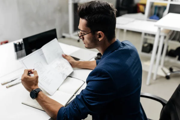 Elegante arquitecto de cabello oscuro en gafas y en una chaqueta azul está trabajando con documentos en el escritorio de la oficina — Foto de Stock