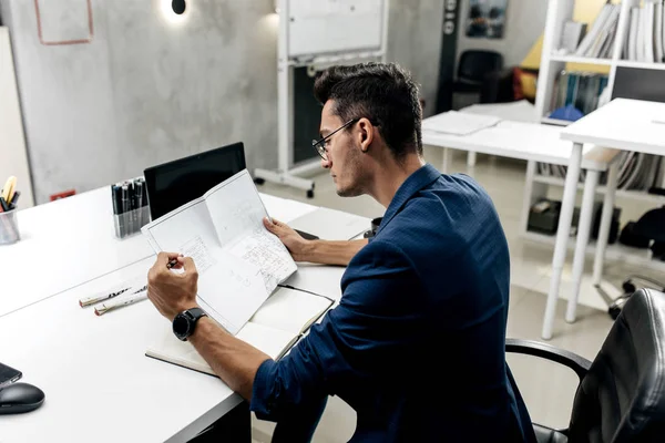 Elegante arquitecto de cabello oscuro en gafas y en una chaqueta azul está trabajando con documentos en el escritorio de la oficina — Foto de Stock