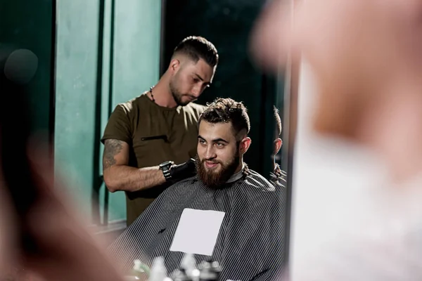 Reflexão no espelho de um barbeiro fazendo uma guarnição de cabelo de jovem homem elegante em uma barbearia — Fotografia de Stock