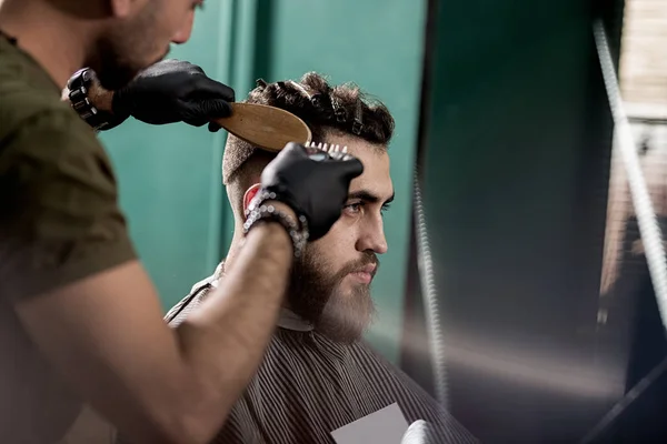 Красивый молодой человек с бородой сидит в парикмахерской. Парикмахер в черных перчатках бреет волосы сбоку . — стоковое фото