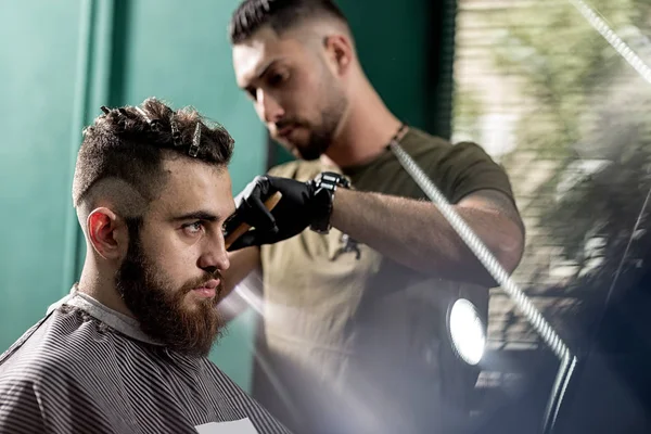 Стильный молодой человек с бородой сидит в парикмахерской. Парикмахер в черных перчатках бреет волосы сбоку . — стоковое фото
