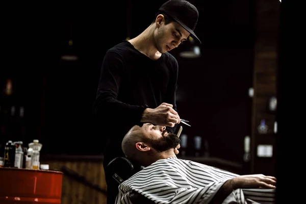 В мужском стиле. Парикмахер одет в черную одежду ножницы бороду жестокого человека в стильной парикмахерской — стоковое фото