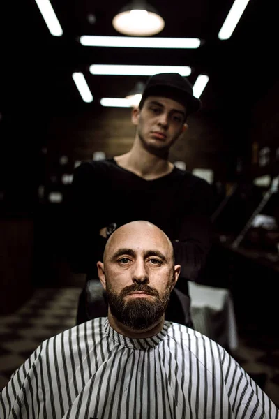 Den snygga barbershop. Mode barberaren står bakom den brutala mannen med skägg sittande i fåtöljen — Stockfoto
