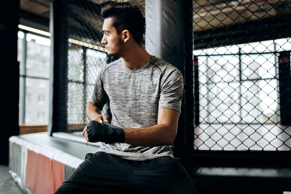Homem de cabelos escuros vestido com a camiseta cinza envolve uma bandagem na mão no ginásio de boxe — Fotografia de Stock