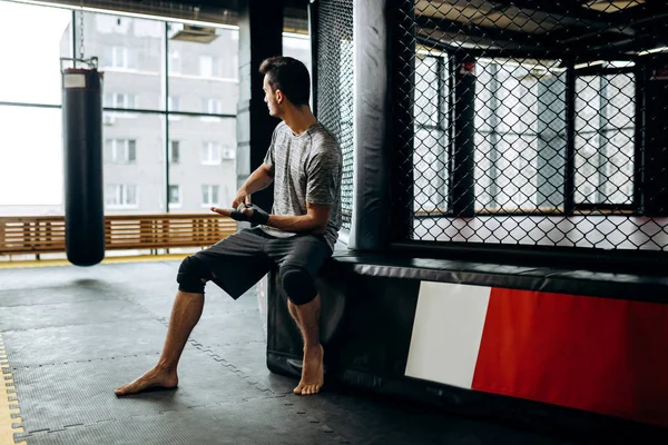 Gri tişört giymiş koyu saçlı adam ve siyah şort boks yüzüğü sınırında oturur ve spor salonunda elinde bir el bandaj sarar — Stok fotoğraf