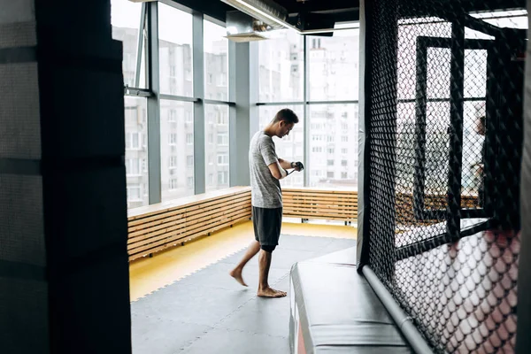 Guy dressed in the grey t-shirt wraps a hand bandage on his hand in the boxing gym against the background of panoramic windows — ストック写真