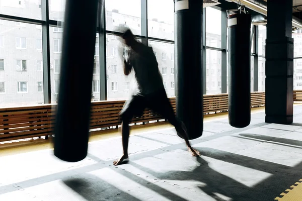 Le gars vêtu du t-shirt gris et du short noir monte la garde et prépare un punch de boxe à côté du sac de boxe suspendu sur le fond des fenêtres panoramiques de la salle de sport — Photo