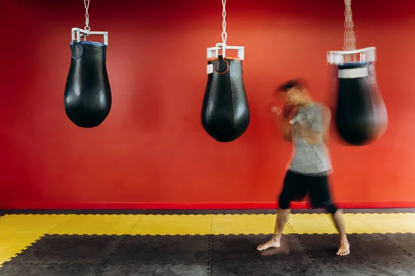 Le mec vêtu du t-shirt gris et du short noir s'entraîne avec un sac de boxe noir contre un mur rouge dans la salle de gym — Photo