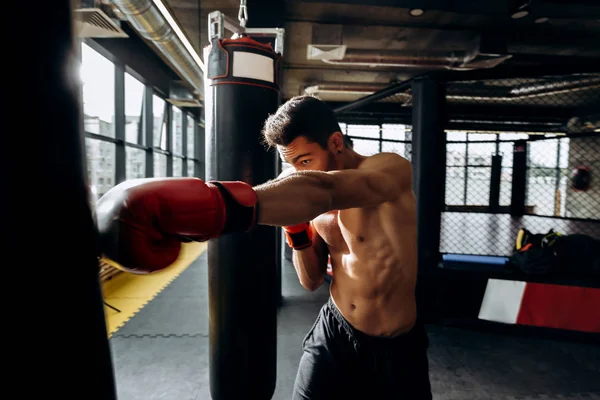 Esportista em luvas de boxe vermelho com um tronco nu bate saco de perfuração no ginásio no fundo do anel de boxe — Fotografia de Stock