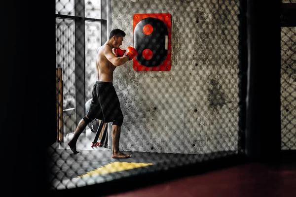 Esportista com um tronco nu e nas luvas de boxe vermelho atinge equipamentos para boxe em uma parede de concreto no ginásio atrás da rede — Fotografia de Stock