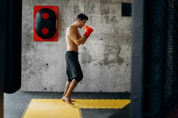 Esportista com um tronco nu e nas luvas de boxe vermelho atinge equipamentos para boxe em uma parede de concreto no ginásio — Fotografia de Stock