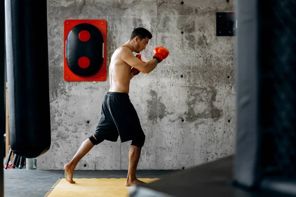 Sportsman avec un torse nu et dans les gants de boxe rouges frappe l'équipement pour la boxe sur un mur en béton dans la salle de gym — Photo