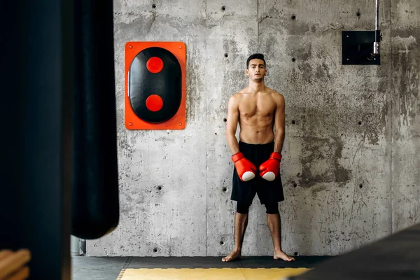 Жорстокий темноволосий хлопець з голим торсом і червоні боксерські рукавички стоять на бетонній стіні поруч з обладнанням для боксу — стокове фото