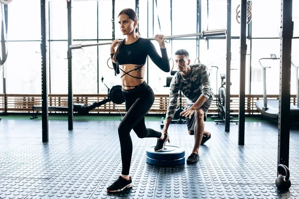 Slim morena chica vestida con ropa deportiva negro está haciendo sentadilla de barra y el entrenador la ayuda en el gimnasio — Foto de Stock