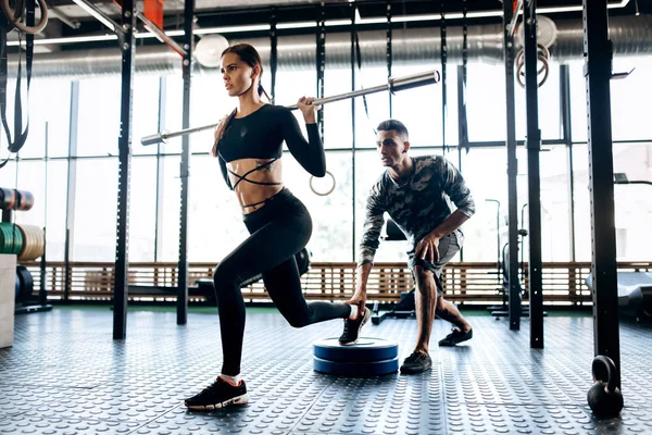 Schlankes dunkelhaariges Mädchen in schwarzer Sportbekleidung macht Langhantel-Kniebeuge und der Trainer hilft ihr im Fitnessstudio — Stockfoto