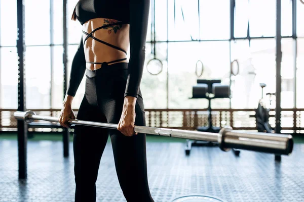 Cuerpo delgado de una chica con tatuaje en una ropa deportiva negra que sostiene en las manos la barra de la barra en el gimnasio — Foto de Stock