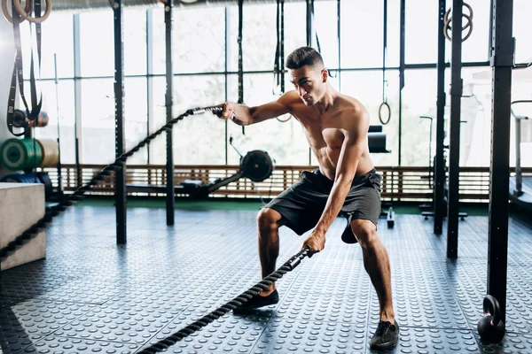 Junger, athletischer Mann mit nacktem Oberkörper in schwarzen Shorts, der in der Turnhalle Sportübungen mit Seilen macht — Stockfoto