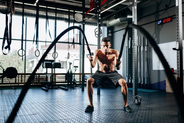 Homme sportif aux cheveux foncés avec un torse nu habillé en short noir faisant des exercices de sport avec des cordes de sport dans la salle de gym — Photo