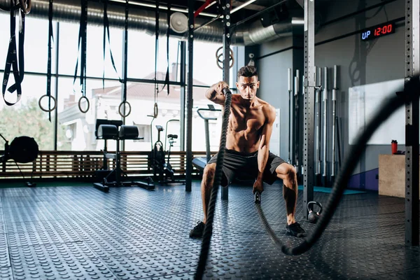 Athletic dark-haired man with a naked torso dressed in the  black shorts doing sport exercises with sports ropes in the gym — Stock Photo, Image