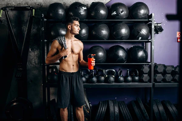 Athletischer dunkelhaariger Mann mit nacktem Oberkörper in schwarzen Shorts steht neben den Sportgeräten in der Turnhalle und hält Flasche mit Wasser in der Hand — Stockfoto