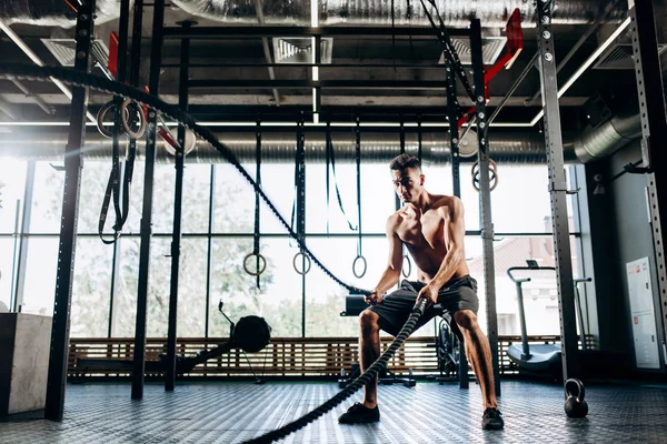 Homem de cabelos escuros atlético com um tronco nu vestido com shorts pretos fazendo exercícios esportivos com cordas esportivas no ginásio — Fotografia de Stock