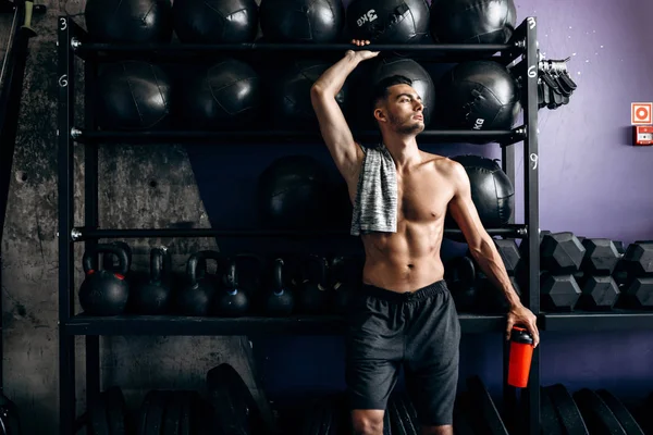 Ein athletischer Mann mit nacktem Oberkörper und T-Shirt auf der Schulter steht in schwarzen Shorts neben den Sportgeräten in der Turnhalle. — Stockfoto