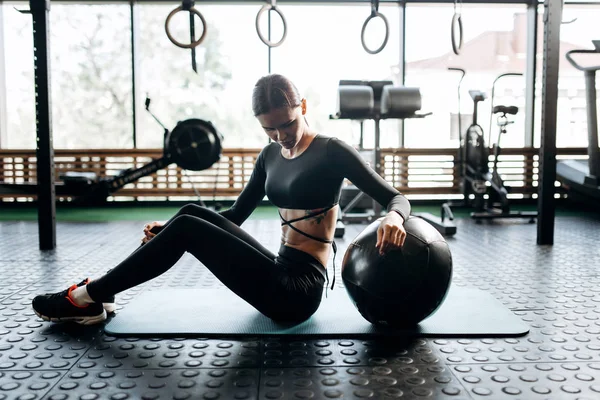 Slim morena chica vestida con ropa deportiva negro está sentado alfombra para la aptitud junto a la pelota de fitness en el gimnasio — Foto de Stock