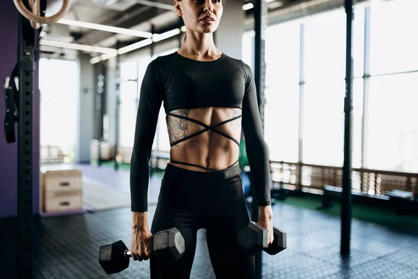 Siyah bir spor giyinmiş dövme olan ince kadın spor salonunda dumbbells ile egzersizleri yapıyor — Stok fotoğraf