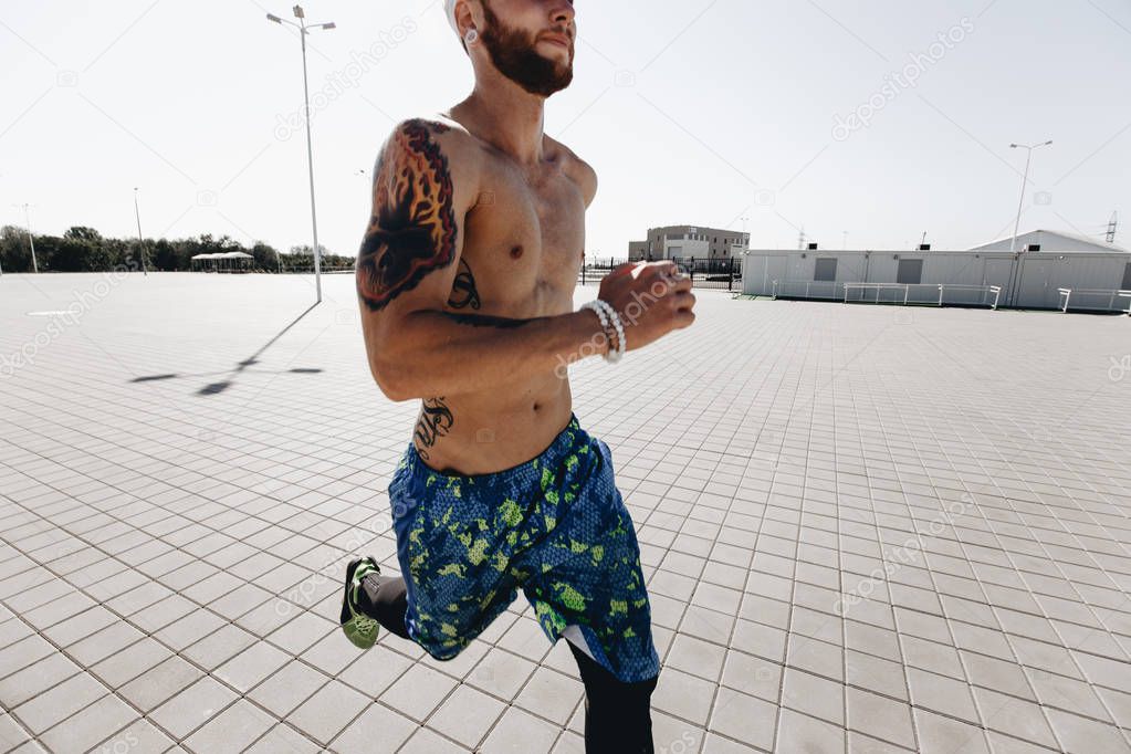 Athletic man with a naked torso with tattoos and with headband dressed in the  black leggings and blue shorts runs on paving slabs on the square on a warm sunny day