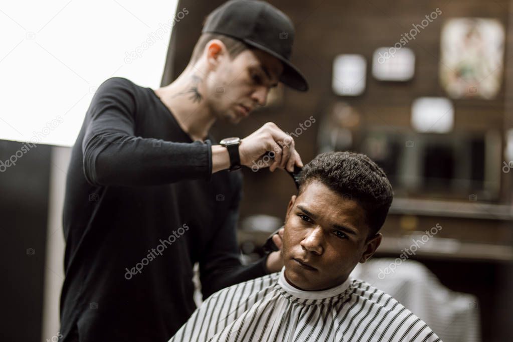 The barber dressed in black clothes  makes a razor cut hair back and sides for a stylish man sitting in the armchair in a barbershop