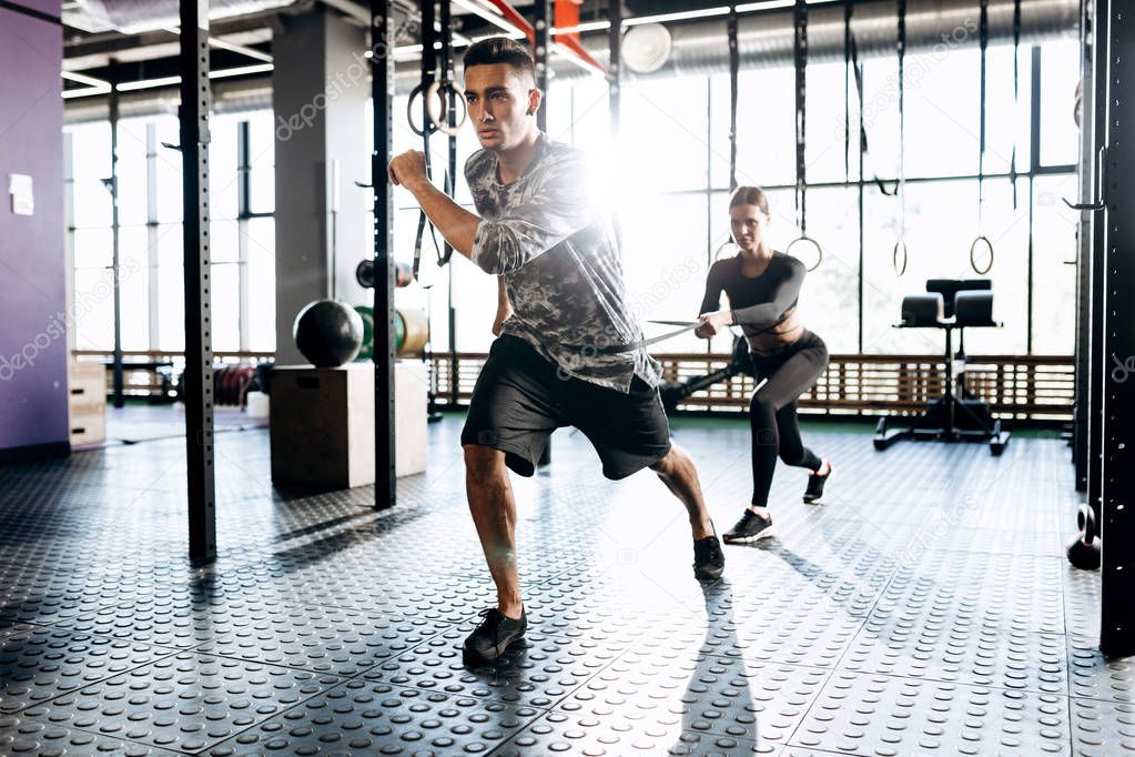 Young athletic man and slim girl are doing sport exercises with special equipment such as power band in the gym